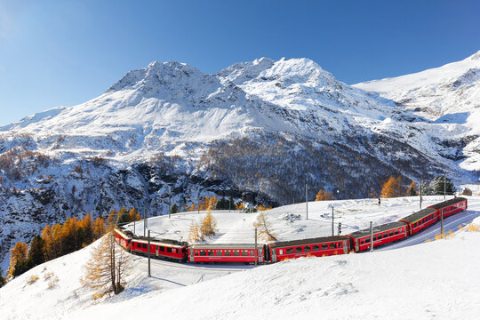 Alp Grum, Switzerland - November 05. 2021 : Red train from Rhaetian Railway is passing the train tracks with tight 180&deg; curve at high Alp Grum. The Piz Palu peak is at the background.