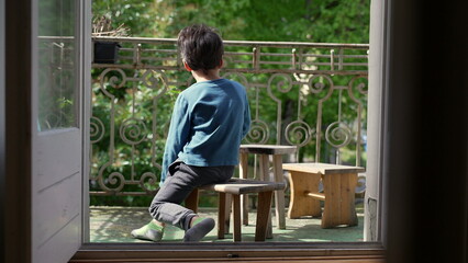 Back view of a boy sitting on a wooden stool on a balcony, looking out into the greenery, a serene...