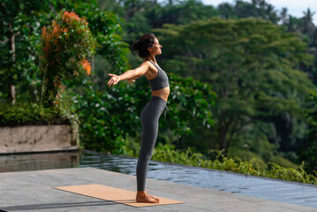 A woman practicing yoga outdoors, standing in a mountain pose with open arms near a tropical pool. Surrounded by lush greenery, she embraces the morning light, promoting wellness, freedom, mindfulness