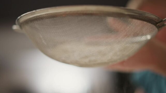 close up hand tapping sieve to sift flour into glass bowl in kitchen setting with soft blurred background showing child silhouette, mixing whisk and rolling pin scattered on counter top