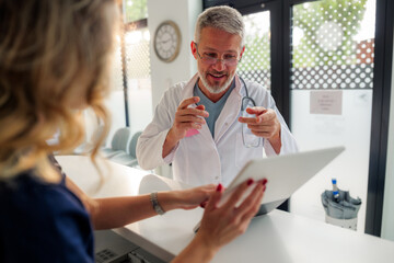 Senior doctor explaining medical information on tablet to nurse at hospital reception desk, discussing patient data and treatment plans in modern healthcare setting