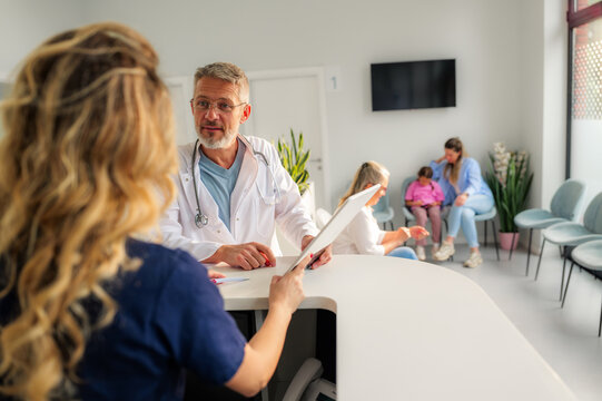 Doctor and nurse are discussing patient information using a tablet at the reception desk of a modern clinic, while patients are waiting in the background
