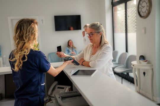 Senior patient using contactless payment for medical services at a modern clinic, interacting with receptionist at the front desk while other patients wait