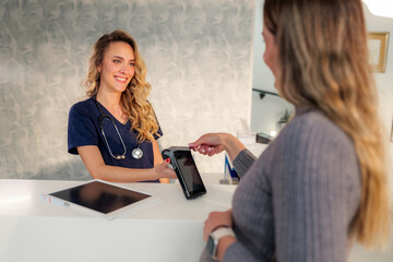 Smiling doctor accepting contactless payment from female patient at modern clinic reception desk,...