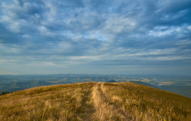 A narrow trail leads over a golden grassy hilltop under a dramatic, cloudy sky, with panoramic views of rolling hills and distant valleys stretching to the horizon.