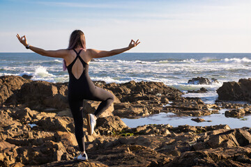 Young woman practicing yoga pose by the ocean on rocky shore, balancing gracefully with open arms under clear blue sky. The scene captures freedom, strength, and a connection with nature.