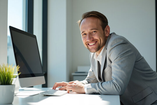 Friendly businessman in a grey blazer pauses from working on his desktop computer to smile warmly at the camera from his desk in a modern office. Approachable professional employee concept