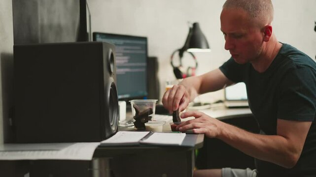 freelancer dipping food into sauce while working on computer with speaker on desk surrounded by containers and notebook in cozy home office environment with warm lighting and blurred background