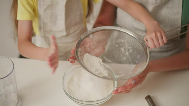 close up of young girl sifting flour into glass bowl while adult supports from behind, teamwork in kitchen surrounded by baking tools and ingredients during cooking session at home