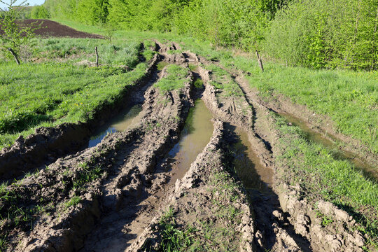 A broken dirt road with deep ruts filled with muddy water runs through a green field. Tracks from heavy machinery are clearly visible on the wet ground, creating an uneven and difficult path to travel