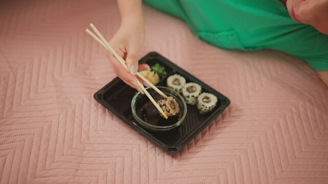 overhead shot of woman in green shirt on pink quilt bed dipping sushi roll into soy sauce with chopsticks slow calm deliberate motion letting excess sauce drip focus on polished nails