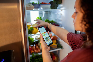 Middle aged mixed ethnicity woman using a dieting app on her smart phone while looking through her fridge