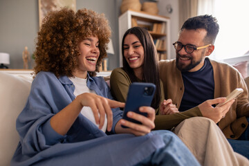 Young Multicultural Friends In Casual Attire Share Entertaining Mobile Content While Relaxing On Comfortable Home Couch