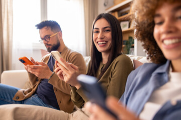 Three Friends Smiling and Chatting on Couch While Using Smartphones in a Cozy Living Room