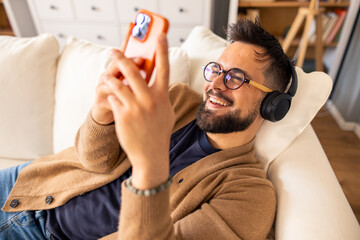 Mid-Adult Man With Headphones And Glasses Smiling While Using Smartphone On Comfortable Living Room...