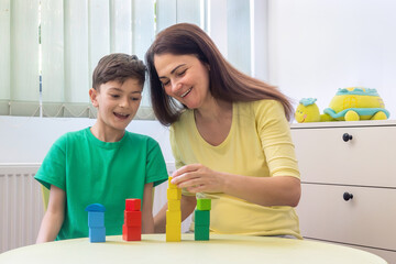 Happy young boy and woman playing with colorful wooden blocks at home. Concept of early childhood education, autism therapy, ADHD support, child psychology, occupational therapy, family connection. 