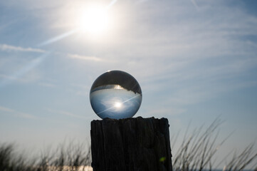 Ball made of glass lies on a wooden post in which the beach and the sea are reflected, dune grasses in the background