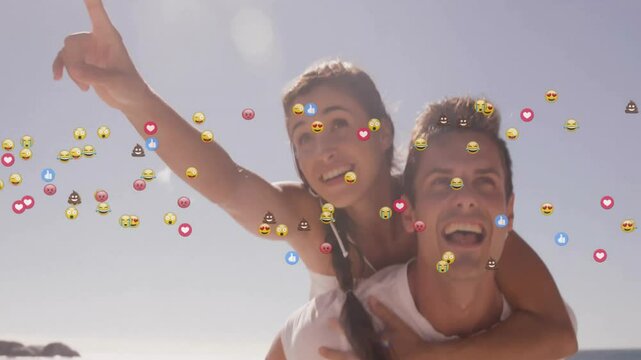 Man giving piggyback ride and woman pointing upward on beach with floating social media emojis