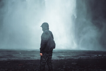 Obraz premium Mann in Outdoor-Kleidung vor Wasserfall im Nebel. Stimmungsvolle Szene eines Mannes in dunkler Outdoor-Kleidung, der an einem nebelverhangenen Wasserfall steht. 