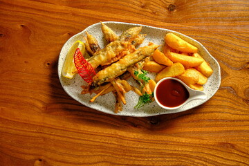 Crispy fish tempura with potato wedges, lemon, greens, and red sauce on a white plate.
