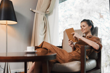 Obraz premium Relaxed young woman in stylish brown outfit enjoying reading on a tablet in a cozy indoor setting with warm lighting