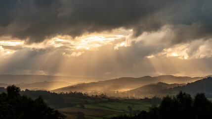 Fototapeta premium Golden Rays Through the Cloudy Sky: Sunbeams pierce through dramatic, overcast clouds, casting a warm, ethereal glow over a rolling landscape of hills and valleys.