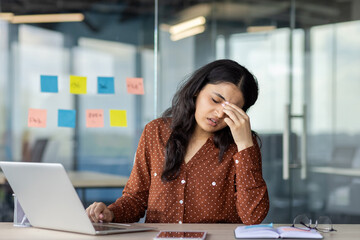 A businesswoman experiencing stress while working on her laptop at her desk in an office setting....
