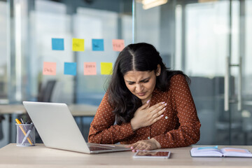 A woman experiencing chest pain sits at her desk, expressing discomfort while working on her...