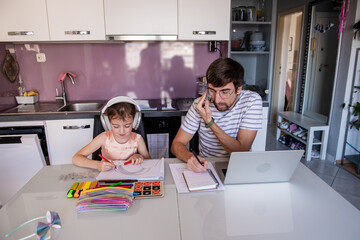 Busy father working from home, taking notes while talking on the phone and his little daughter drawing and wearing headphones in the kitchen