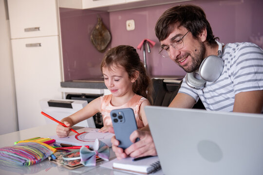 Father wearing headphones, working on his laptop while capturing a picture of his daughter's colorful drawing with a smartphone in a cozy home office setup in the kitchen - Powered by Adobe
