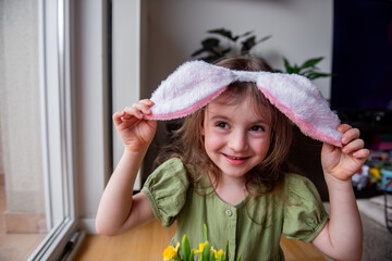 Little girl wearing festive Easter bunny ears, joyfully holding bright daffodils and smiling at...