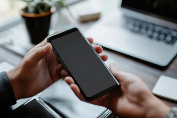 Person holding a smartphone with a blank screen in front of a laptop on a wooden desk surface