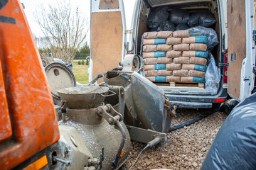 Outdoor construction site featuring a van with open rear doors, cement bags on a pallet, a concrete...
