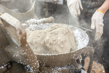 Powdered material and white granules mix in a metal container, with gloved hands nearby. Dust particles are visible, indicating an industrial process.