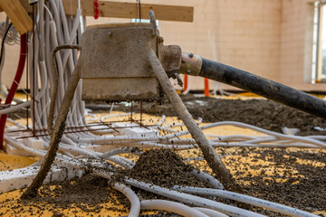 Concrete pouring machine dispenses material on a yellow base layer with rebar. Scattered cables, pipes, and unfinished walls indicate early construction work.