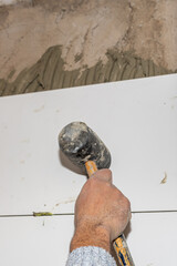 Construction worker is using a rubber mallet to place tiles on a wall, installing large format white tiles