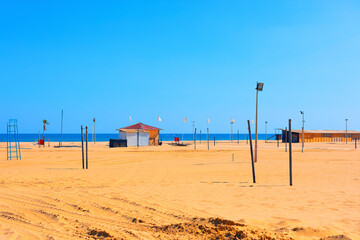 Wide sandy beach in Catania stretches under a clear sky, with calm sea in background and a lifeguard station near horizon. Volleyball nets, lifeguard tower, and scattered poles dot empty shoreline