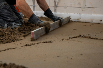 A worker kneels on wet concrete, leveling it with a straightedge tool. Underfloor heating pipes are visible, with unfinished walls and wiring around.