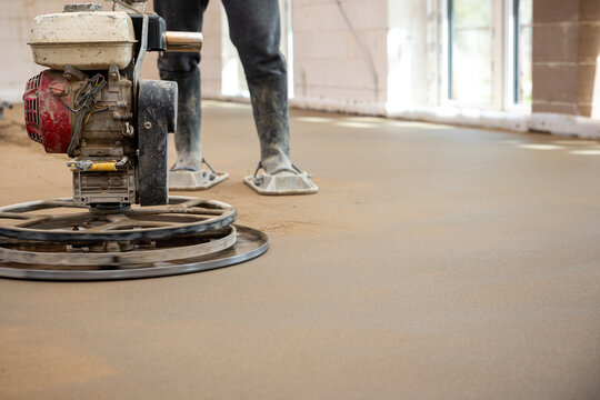 A construction worker smooths screed concrete with a power trowel, wearing protective footwear. Underfloor heating pipes are installed beneath the surface.