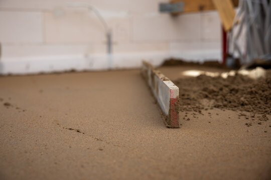 A screed tool levels freshly laid Estrich concrete indoors, with white walls, electrical outlets, and cables visible in the background.