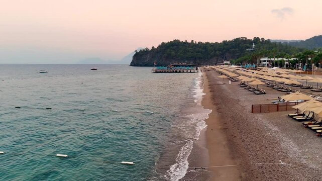 Colorful evening scene of a peaceful beach with rows of sunbeds and umbrellas, gentle waves along the shore, and a scenic pier under a vibrant sky. 