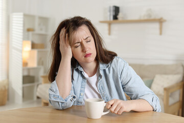 Suffering from hangover. Unhappy woman with cup of coffee sitting at wooden table indoors
