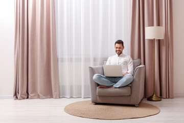 Smiling man holding laptop on armchair near window with beautiful curtains at home. Space for text