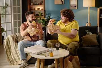 Two friends eating fast food burgers and smiling in cozy living room with wooden elements and modern decor, enjoying relaxed and cheerful moment together