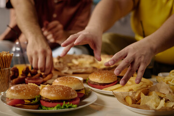 Group of people reaching for burgers and chips on a table indoors, enjoying casual time together. Food spread includes burgers, chips, and other snacks with drinks visible in background