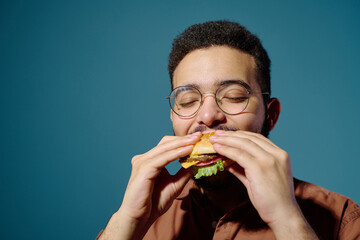 Young man enjoying his burger with eyes closed, savoring every bite, standing against a plain blue...