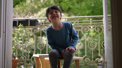 Young boy sitting on a wooden stool on a balcony, smiling and looking up, surrounded by lush...