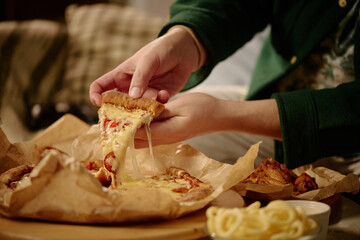 Person holding and pulling slice of melted cheese pizza from fresh-baked whole pizza. Background includes cozy home setup with blurred furniture