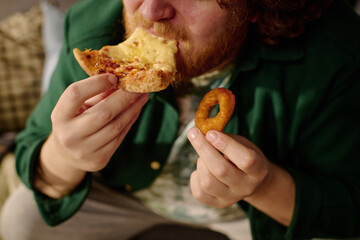Caucasian man enjoying a slice of pizza and holding an onion ring at home. Wearing casual attire...