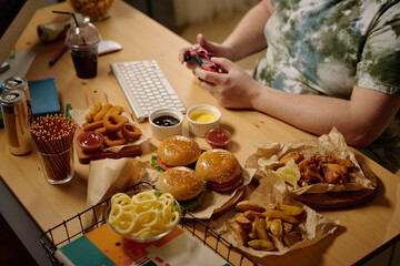 Person enjoying gaming while surrounded by an assortment of snacks, including burgers, fries, onion rings, and soda cans. Gamer holding a controller with a focused expression